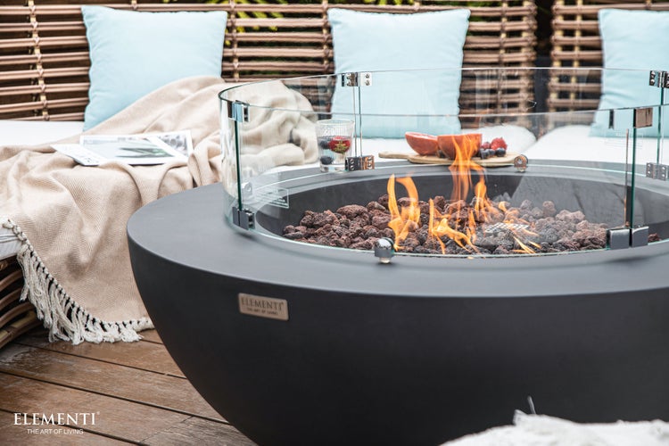 Runder schwarzer Gas-Feuertisch mit Glas-Windschutz und Lavasteinen auf einer Holzterrasse neben einem Sofa mit Kissen. Elementi Logo.