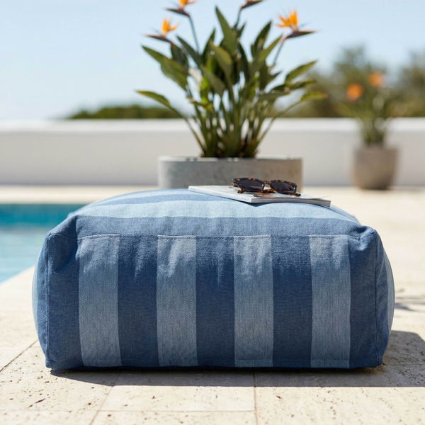 Rechteckiger Outdoor-Pouf mit blauen Längsstreifen auf einer Terrasse am Pool mit einer Zeitschrift und Sonnenbrille darauf.