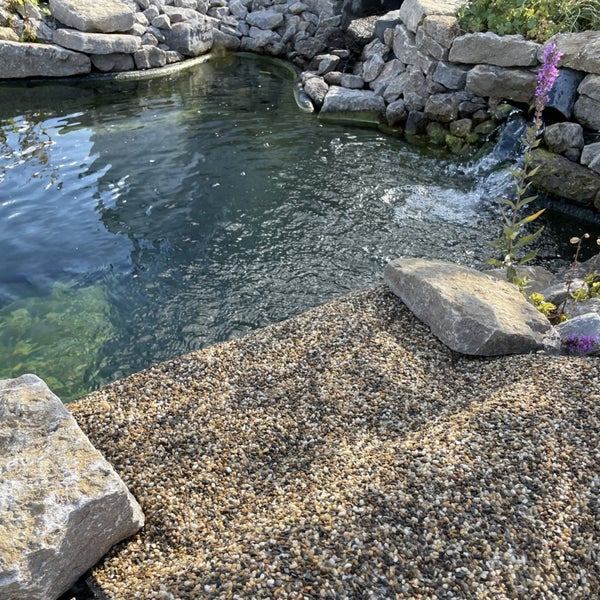 Ein angelegter Gartenteich mit einem kleinen Wasserfall, umrandet von Natursteinen und einer Kiesfläche im Vordergrund.