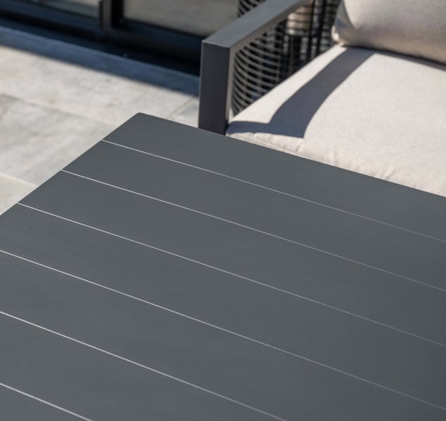 Detailansicht eines anthrazitfarbenen Gartentischs mit Lattenstruktur neben einem Loungesessel mit hellen Kissen auf einer Terrasse.