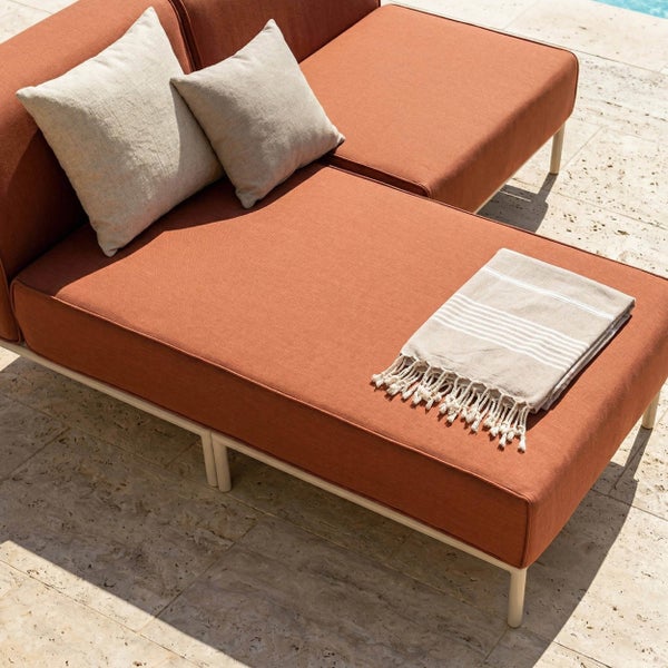 Modernes Garten-Daybed mit rostfarbenen Polstern, beigen Kissen und einem gestreiften Handtuch auf einer Steinterrasse.
