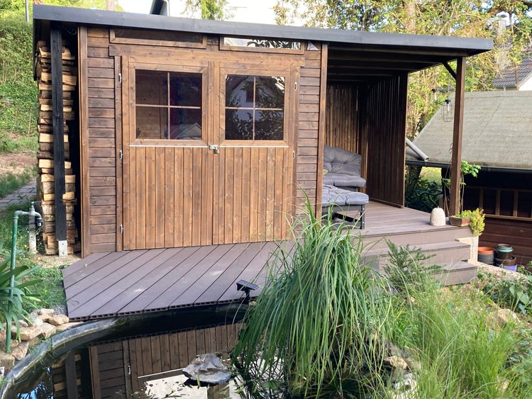 Modernes Gartenhaus aus Holz mit Doppeltür, Fenstern, integrierter Terrasse und Kaminholzregal an einem Gartenteich.