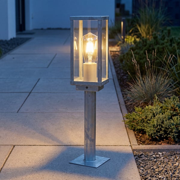 Sockelleuchte für den Außenbereich aus verzinktem Stahl mit Glaseinsätzen und leuchtendem Leuchtmittel auf einem Gartenweg.
