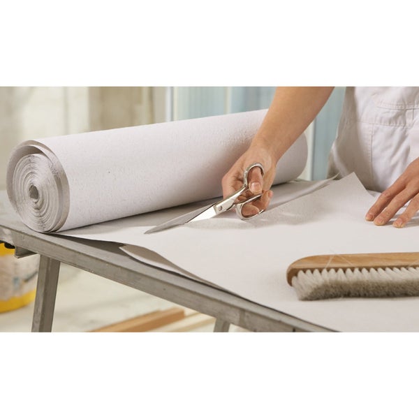 Eine Person schneidet eine Rolle weiße Tapete mit einer Tapetenschere auf einem Tapeziertisch neben einer Tapezierbürste zu.