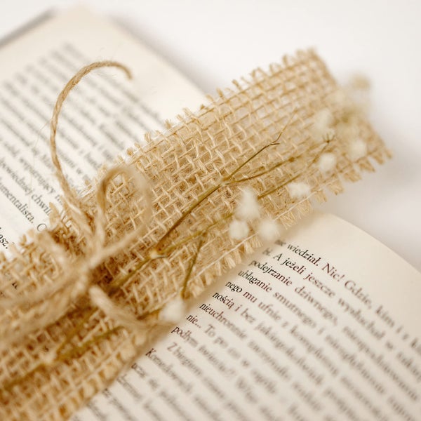 Dekoratives Lesezeichen aus Jutegewebe mit Kordelschleife und Trockenblumen, auf einem aufgeschlagenen Buch liegend.