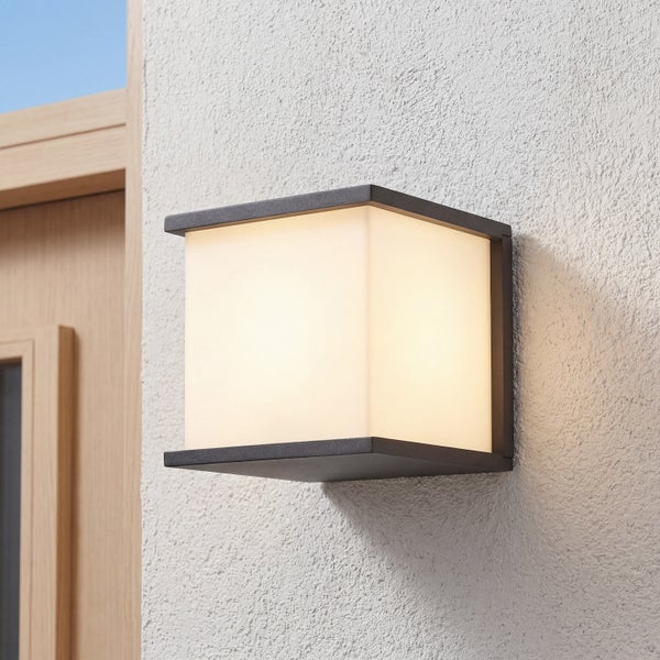 Moderne, quadratische Außenwandleuchte an einer weißen Putzwand neben einem Türrahmen aus Holz.