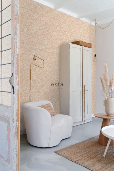 Vliestapete mit floralem Muster in hellem Wohnraum mit Polstersessel, Holzschrank und Beistelltisch. esta home Logo.