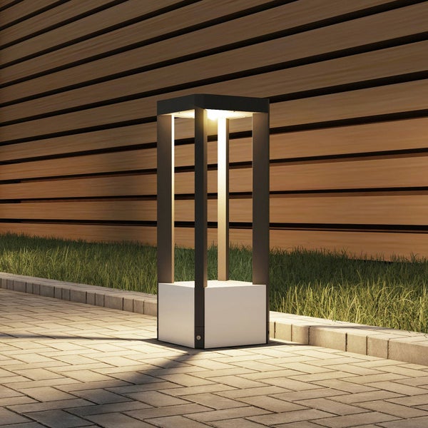 Moderne Außen-Sockelleuchte auf einem gepflasterten Weg neben einer Rasenfläche und einer Holzwand, hell beleuchtet.