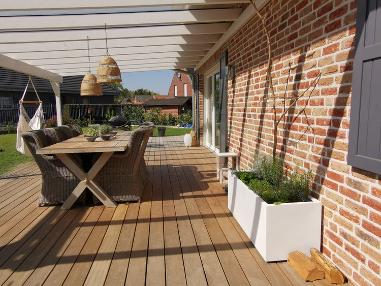 Moderne Terrasse mit Holzdielen, Backsteinwand, weißem Pflanzkasten, Holztisch und Rattanstühlen unter einer weißen Überdachung.