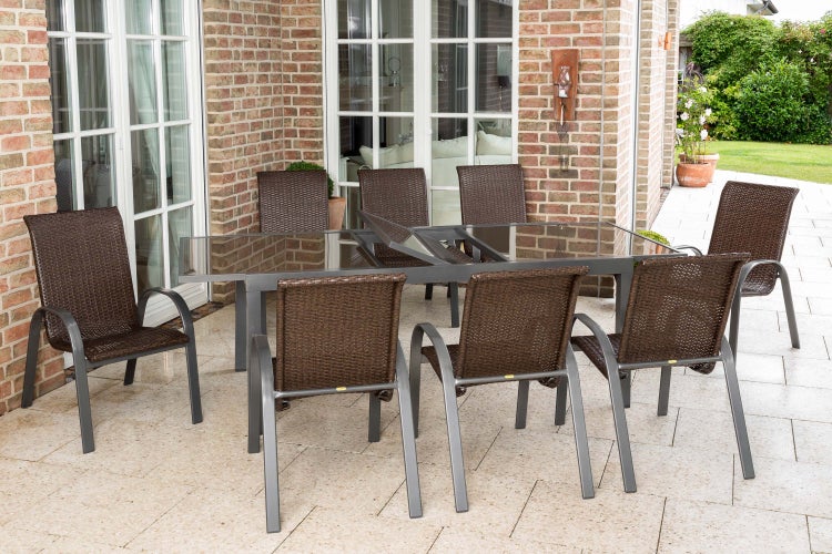 Gartenmöbel-Set mit ausziehbarem Esstisch mit Glasplatte und acht Stapelstühlen aus Polyrattan auf einer Terrasse vor einer Ziegelwand.