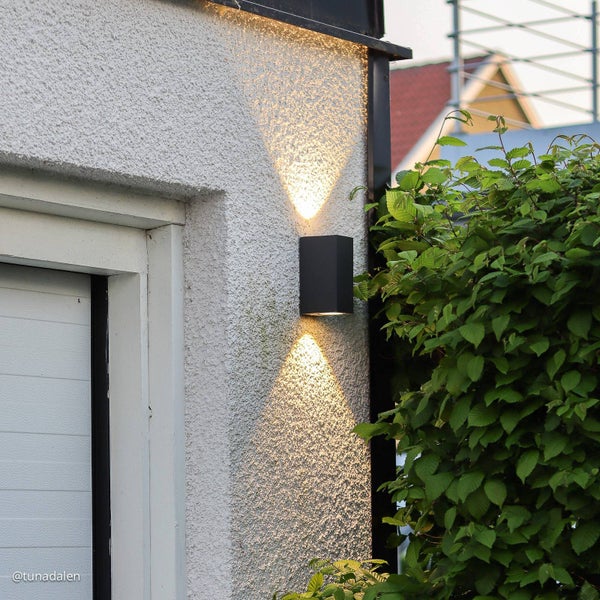 Moderne schwarze rechteckige Außenwandleuchte mit Lichtaustritt nach oben und unten an einer weißen Fassade neben grünen Pflanzen.