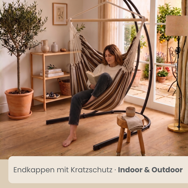 Hängesessel mit Gestell für Innen und Außen, Frau liest in modernem Wohnbereich vor einer Terrasse mit bodentiefen Fenstern.