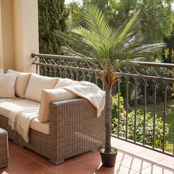 Künstliche Palme in einem schwarzen Topf neben einem Loungesofa mit hellen Polstern auf einem überdachten Balkon mit Metallgeländer.