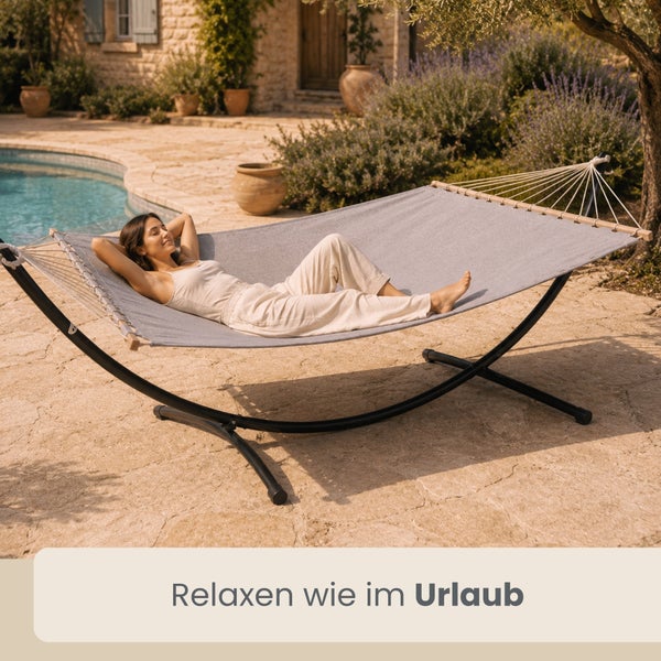 Eine Frau entspannt in einer Stoffhängematte mit Hängemattengestell und Spreizstäben auf einer Steinterrasse neben einem Swimmingpool.