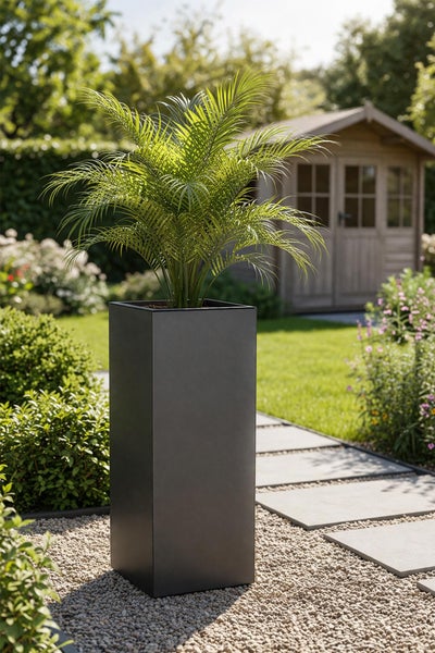 Hoher rechteckiger Pflanzkübel in Anthrazit mit einer Palme auf einem Kiesweg in einem Garten mit Gartenhaus und Rasen.