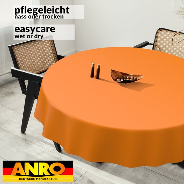 Rundes, orangefarbenes, pflegeleichtes Tischtuch auf einem Tisch mit zwei Rattanstühlen. ANRO Deutsche Manufaktur Logo.