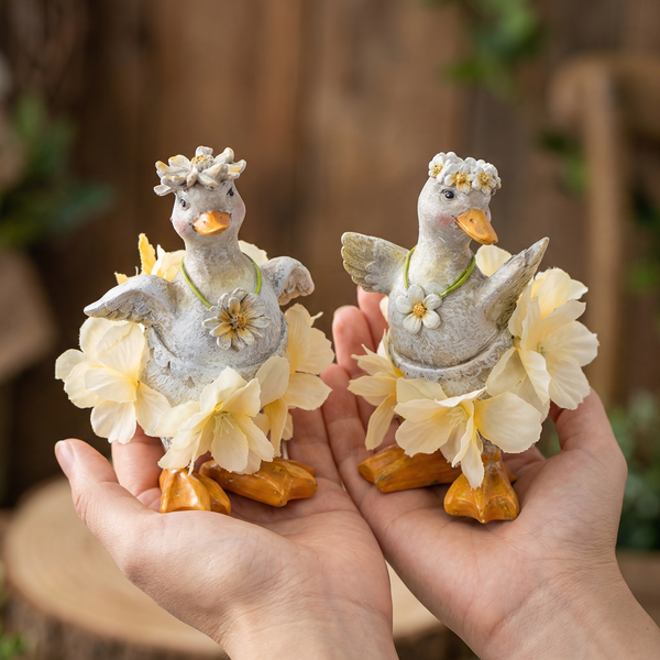 Zwei dekorative Entenfiguren mit Blumenkränzen und gelben Stoffblumen, in den Händen gehalten.