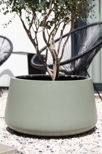 Moderner salbeigrüner Pflanzkübel mit einer Pflanze auf einer Kiesterrasse, im Hintergrund schwarze Gartenstühle.