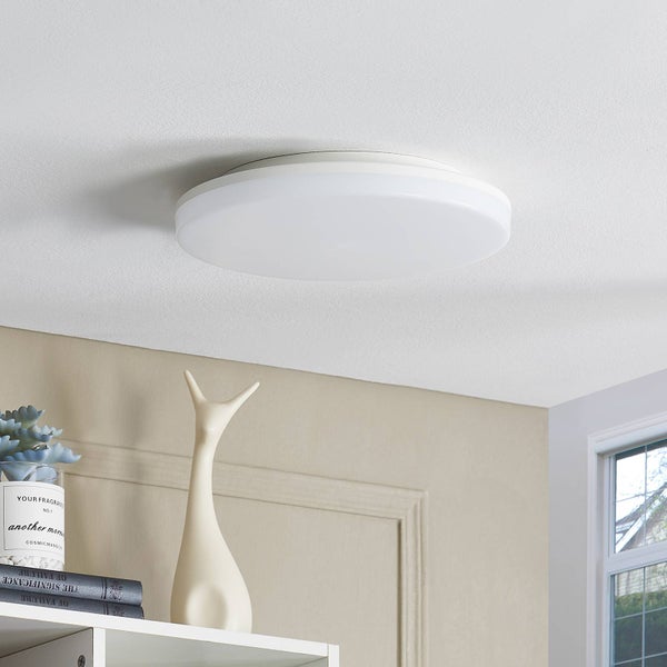 Moderne runde weiße LED Deckenleuchte montiert an der Decke über einem Regal mit Dekoration in einem hellen, ordentlichen Raum.