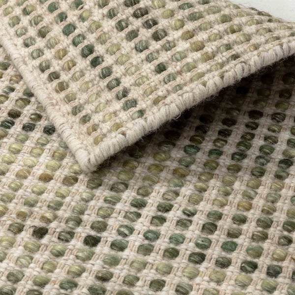 Nahaufnahme eines Webteppichs mit grünen Filzkugeln in einem beigen Gitter, die strukturierte Oberfläche und eine umgeschlagene Ecke zeigend