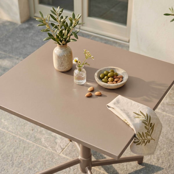 Quadratischer Gartentisch aus Metall auf einer Terrasse, dekoriert mit einer Vase mit Olivenzweigen, einer Schale mit Oliven und einer Stoffserviette.