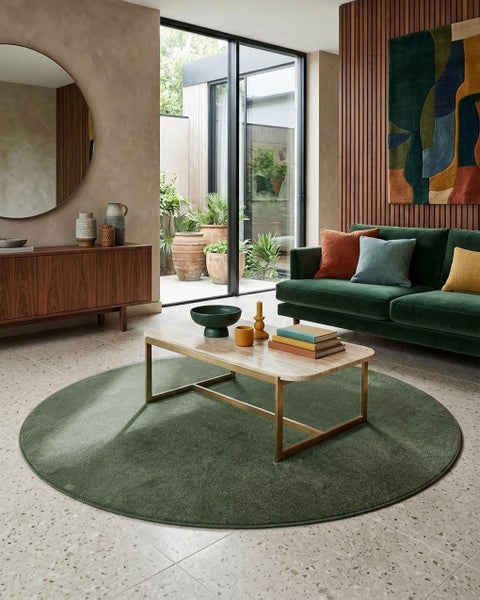 Modernes Wohnzimmer mit rundem grünem Teppich, Marmor-Couchtisch, grünem Samtsofa und Sideboard aus Holz vor einer Glasfront.