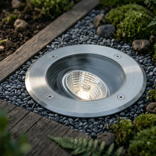 Runder Bodeneinbaustrahler aus Edelstahl installiert in einem Garten zwischen dunklem Kies und Moos.