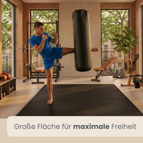 Heller Fitnessraum mit einem Mann auf großen schwarzen Bodenschutzmatten, Boxsack und verschiedenen Fitnessgeräten.