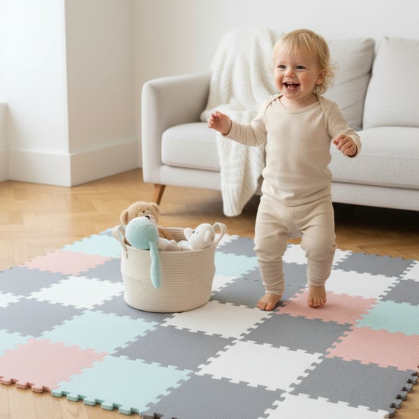 Kleinkind steht auf einer bunten Puzzlematte aus Schaumstoff in einem hellen Wohnzimmer neben einem Korb mit Stofftieren.
