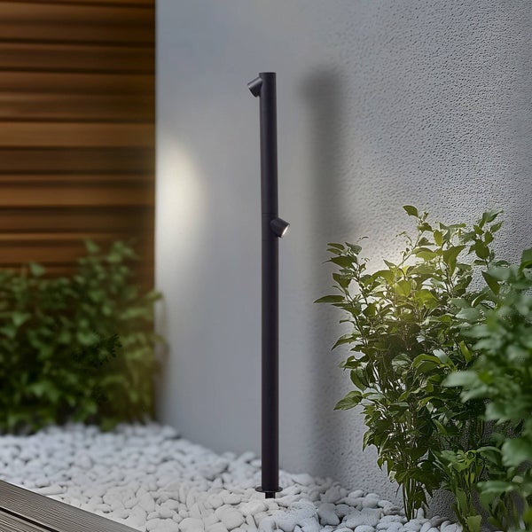 Schlanke schwarze Pollerleuchte mit zwei Strahlern, die eine strukturierte Wand und Pflanzen in einem hellen, organisierten Garten beleuchten.