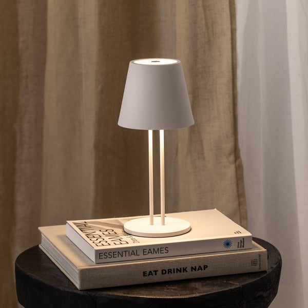 Moderne Tischleuchte mit kegelförmigem Schirm und schlankem Doppelstabgestell auf einem Stapel Bücher in warmem Licht vor einem Vorhang.