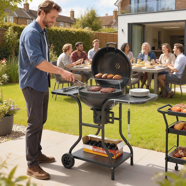 Ein Mann grillt Fleisch und Kartoffeln auf einem schwarzen Holzkohle-Kugelgrill in einem Garten, während Gäste an einem Tisch sitzen.