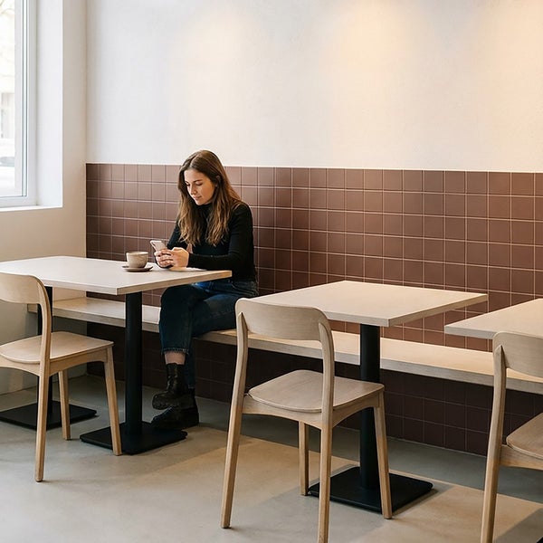 Eine Frau sitzt an einem Tisch in einem modernen Raum mit quadratischen Wandfliesen und hellen Holzstühlen.