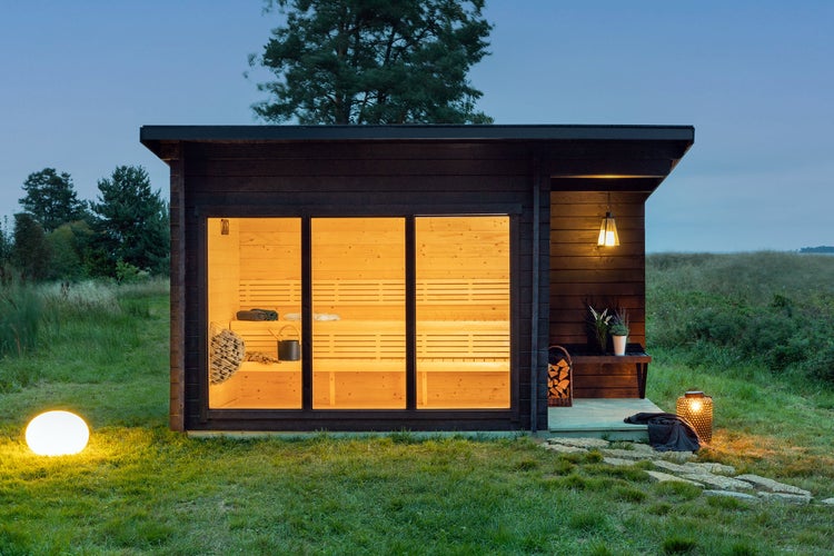 Modernes Gartensaunahaus mit Flachdach, dunkler Holzfassade und großen Glasfronten, hell beleuchteter Innenraum mit Holzbänken am Abend.