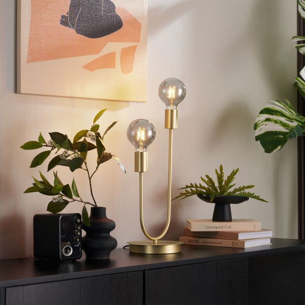 Moderne Tischleuchte aus Messing mit zwei U-förmigen Armen und sichtbaren Leuchtmitteln auf einem schwarzen Sideboard mit Dekoration.