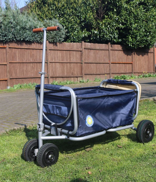 Zusammenklappbarer Bollerwagen mit Metallgestell, Textilbespannung und Luftreifen auf einer Wiese vor einem Holzzaun.