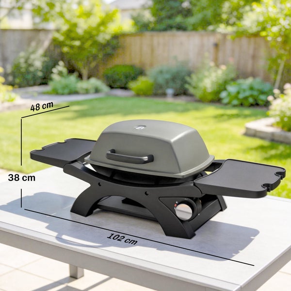 Grauer tragbarer Gasgrill auf einem Tisch im hellen Garten mit Maßen 102 Zentimeter Breite, 38 Zentimeter Höhe und 48 Zentimeter Tiefe.