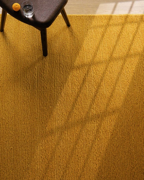 Strukturierter gelber Teppich mit Rippenstruktur. Ein Hocker mit einem Glas Wasser und einer Orange steht auf dem Teppich.