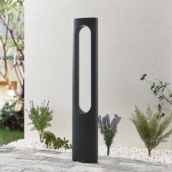 Moderne anthrazitfarbene Wegeleuchte aus Metall mit ovalem Ausschnitt in einem Garten mit Pflanzen und Kieselsteinen.