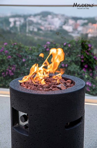 Modeno runder Gaskamin in schwarzer Beton-Optik mit Lavasteinen und brennenden Flammen auf einer Terrasse. Modeno Logo.