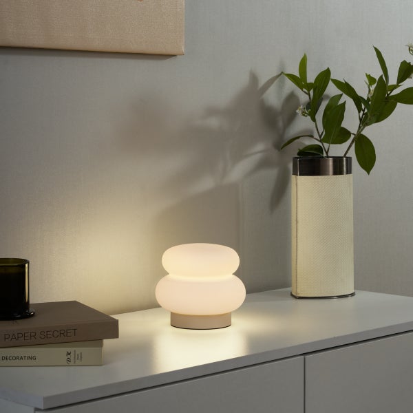 Moderne, zweistufige Tischleuchte aus Glas mit warmem Licht auf einem weißen Sideboard neben Büchern, einer Kerze und einer Vase.