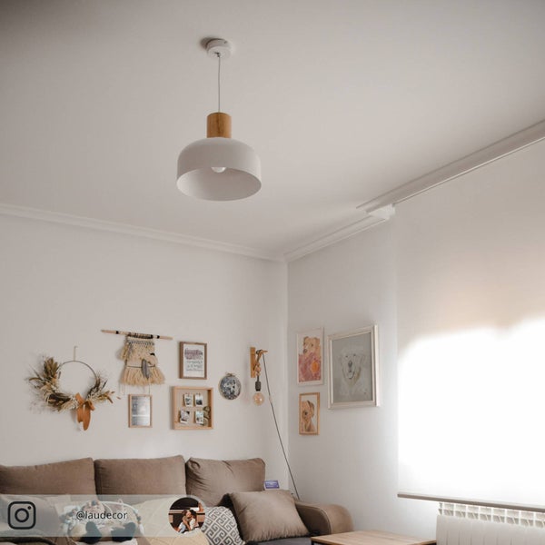 Helle Wohnzimmerszene mit weißer Pendelleuchte aus Metall und Holz über einem braunen Sofa vor einer dekorierten Wand.