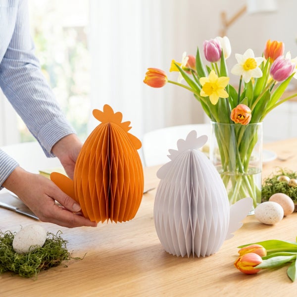Dekorative Wabenpapierhühner als Osterdekoration auf einem Holztisch mit Blumenstrauß und Ostereiern.