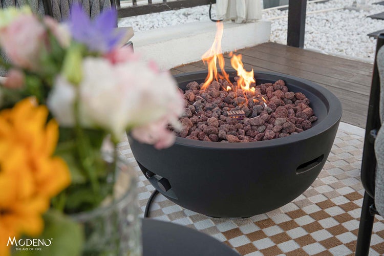 Runde Gas-Feuerschale in Anthrazit mit Lavasteinen und Flammen auf einer Terrasse mit gemustertem Außenteppich. Modeno Logo.