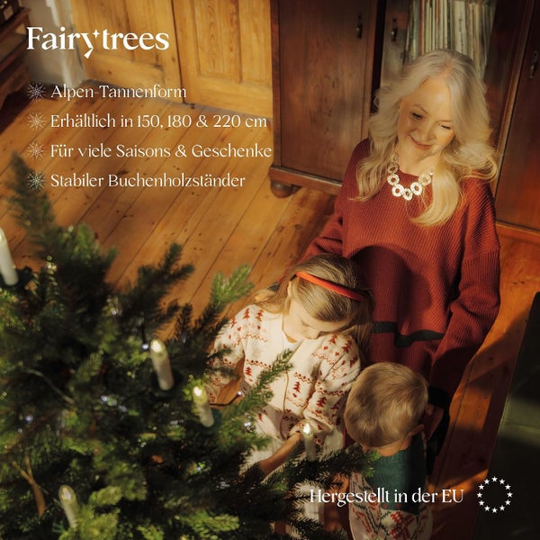 Familie schmückt einen Fairytrees Kunstweihnachtsbaum im Alpen Tannen Design mit Buchenholzständer in einem gemütlichen Wohnzimmer.