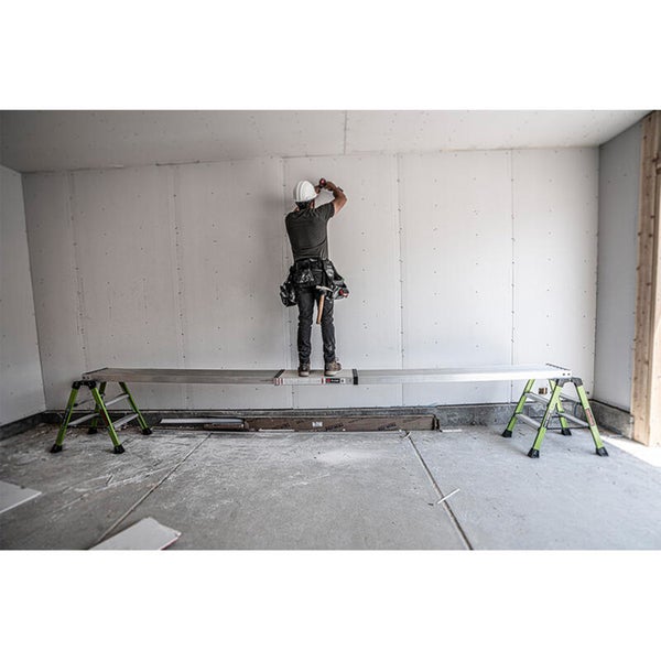 Handwerker arbeitet auf einem langen Aluminium Arbeitsgerüst vor einer Trockenbauwand in einem unfertigen Innenraum