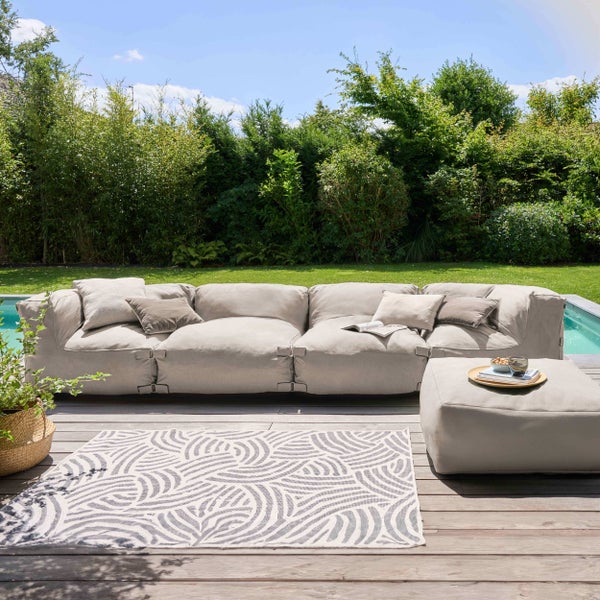 Modulares Outdoor Lounge Sofa mit Kissen, Hocker und gemustertem Außenteppich auf einer Holzterrasse am Pool im hellen Garten.