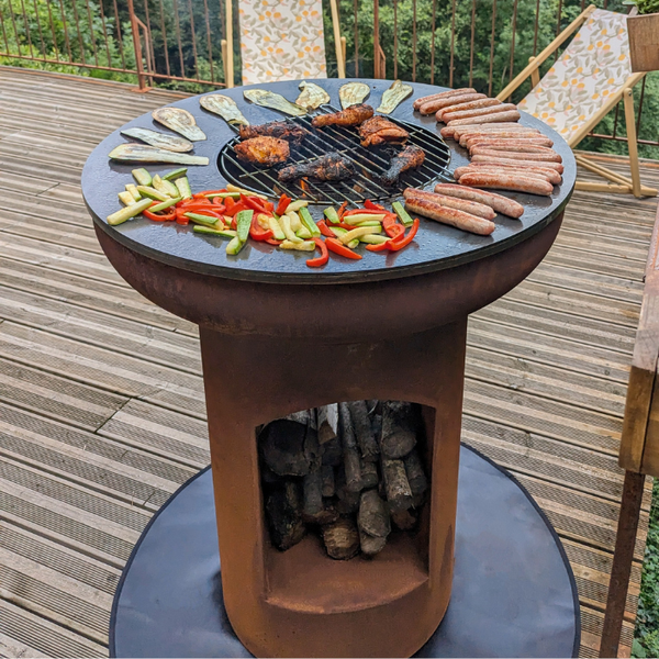 Runder Plancha-Grill aus Cortenstahl mit integriertem Holzfach auf einer Holzterrasse, belegt mit Fleisch und Gemüse.
