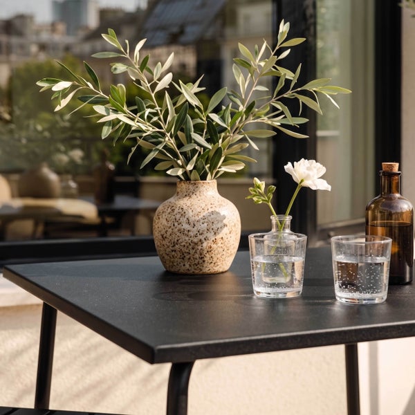 Schwarzer Metall-Gartentisch dekoriert mit einer Keramikvase mit Olivenzweigen, einer Blume und Gläsern auf einer hellen Terrasse.