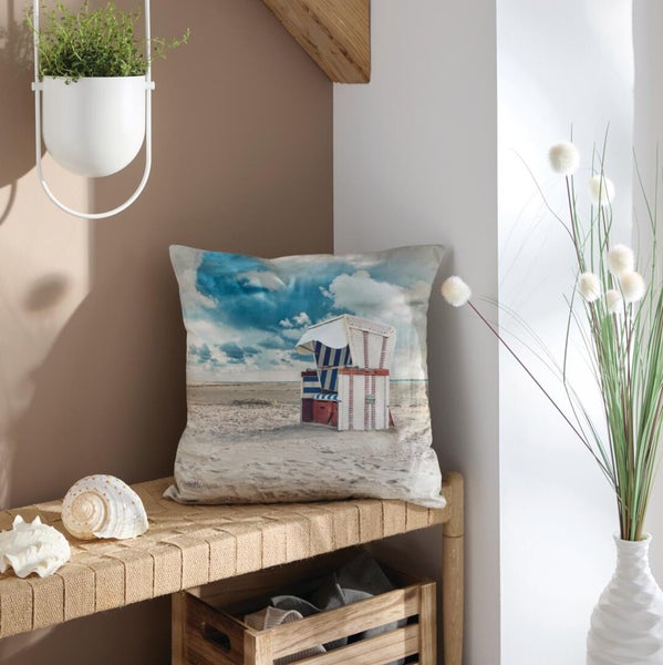 Dekokissen mit Strandkorb-Motiv auf einem Sandstrand unter blauem Himmel, platziert auf einer geflochtenen Bank mit Muscheln.